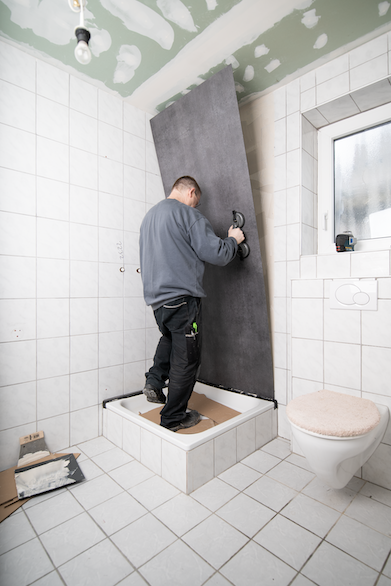 Ein Mann renoviert ein Bad und bringt eine großes, graues Ceramin Tiles Element an der Wand hinter der Dusche an.
